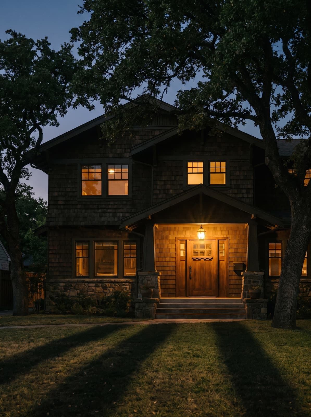 A craftsman house at dusk under oak trees, porch light on